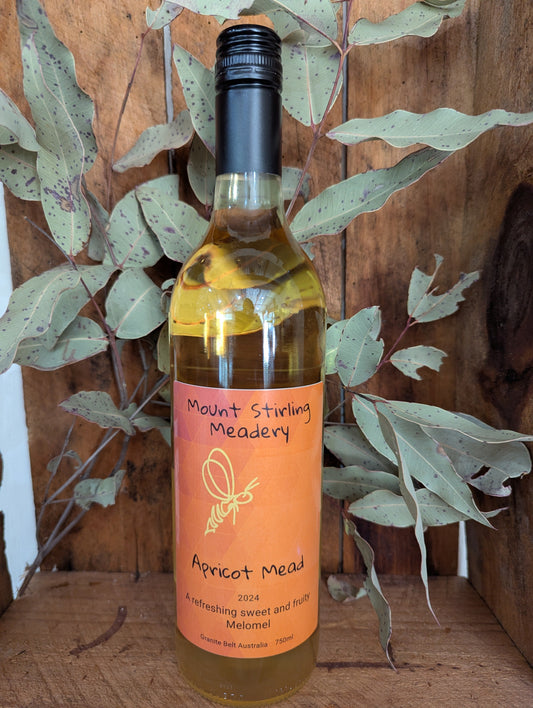 A bottle of Apricot Melomel Mead by Mount Stirling Meadery, with a yellow label and a dark cap, placed against a wooden background with green foliage.